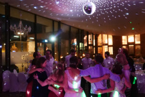 Eine Gruppe von Hochzeitsgästen in festlicher Kleidung steht im Kreis, sie tanzen und umarmen sich. Sie genießen einen stimmungsvollen Abend unter einer schimmernden Discokugel. Der Raum ist schwach beleuchtet, verstreute Lichtmuster an Decke und Wänden tragen zur festlichen Atmosphäre bei. DJ Martin Meyer ist für den schönsten Tag in Ihrem Leben verantwortlich!