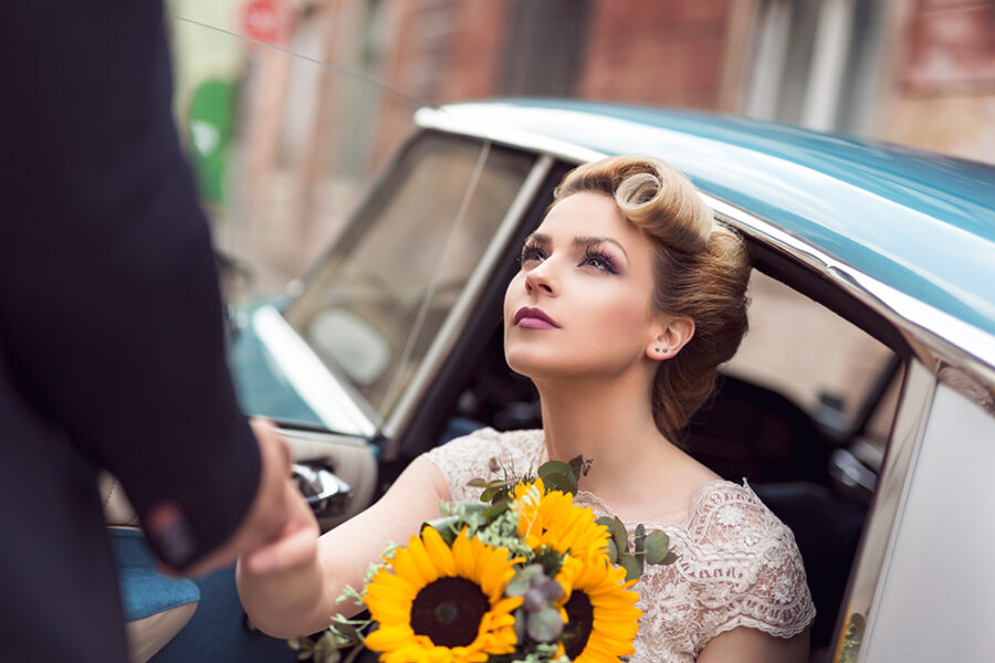 Beautiful young bride sitting in a wedding dress in a retro old car, holding a sunflower bouquet while groom is helping her get out of the car