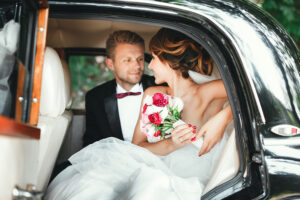 Wedding day. Bride and groom sitting in wedding car, looking at each other, smiling and holding bouquet. Decorations. Opened door