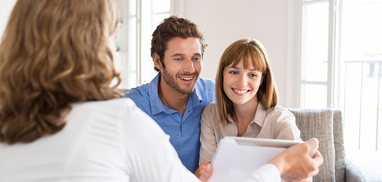 Ein Paar sitzt zusammen auf einem Sofa, lächelt und schaut auf ein Tablet, das eine Person mit dem Rücken zur Kamera hält. Der Raum ist hell, natürliches Licht fällt durch große Fenster.