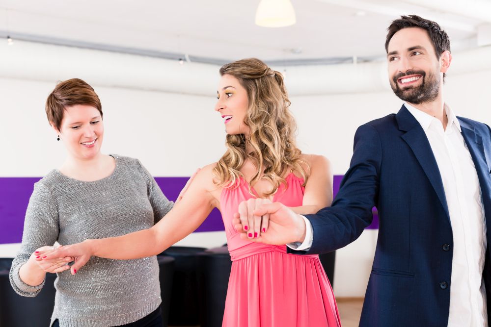 Drei Menschen lächeln und halten sich an den Händen, während sie drinnen tanzen: eine Frau in einem grauen Pullover, eine Frau in einem rosa Kleid und ein Mann im Anzug. Die Szene fängt die Freude des Hochzeitstanzes in einem hell erleuchteten Raum mit einem violetten Streifen an der Wand ein.