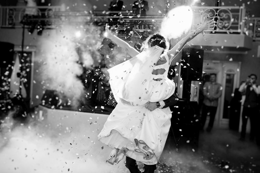 Braut und Bräutigam umarmen sich während des Hochzeitstanzes, umgeben von Konfettiregen. Das monochrome Bild fängt die festliche Atmosphäre ein, im Hintergrund sind Gäste und kunstvolle Dekorationen zu sehen.