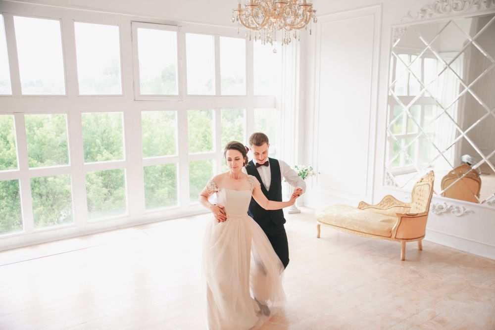 Braut und Bräutigam gleiten durch „Der Hochzeitstanz“ in einem hellen, eleganten Raum mit großen Fenstern. Die Braut trägt ein fließendes weißes Kleid und der Bräutigam einen schwarzen Smoking. Über ihnen funkelt ein Kronleuchter, während sie sich anmutig neben einem Vintage-Sofa an der Wand bewegen.