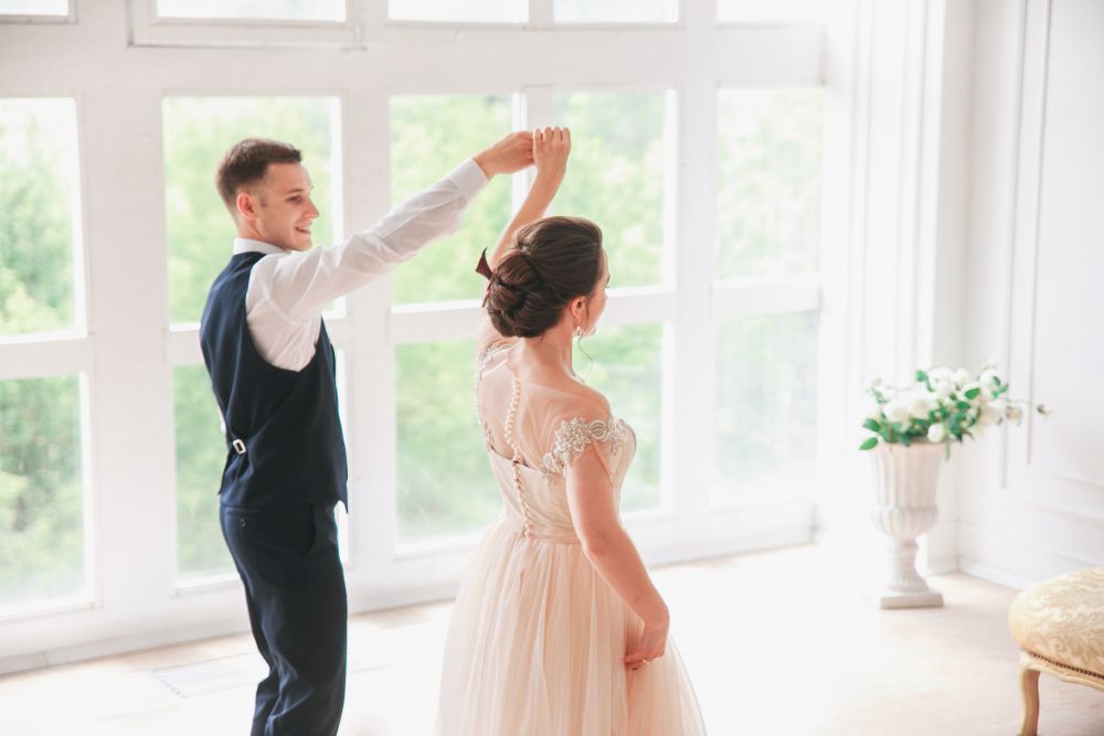 Der Hochzeitstanz entfaltet sich, während der Bräutigam die Braut anmutig in einem hellen Raum wirbelt. Sie glänzt in einem eleganten Kleid mit Spitzendetails, während er eine dunkle Weste und Hose trägt. Natürliches Licht strömt durch ein großes Fenster und hebt ein Blumenarrangement im Hintergrund hervor.
