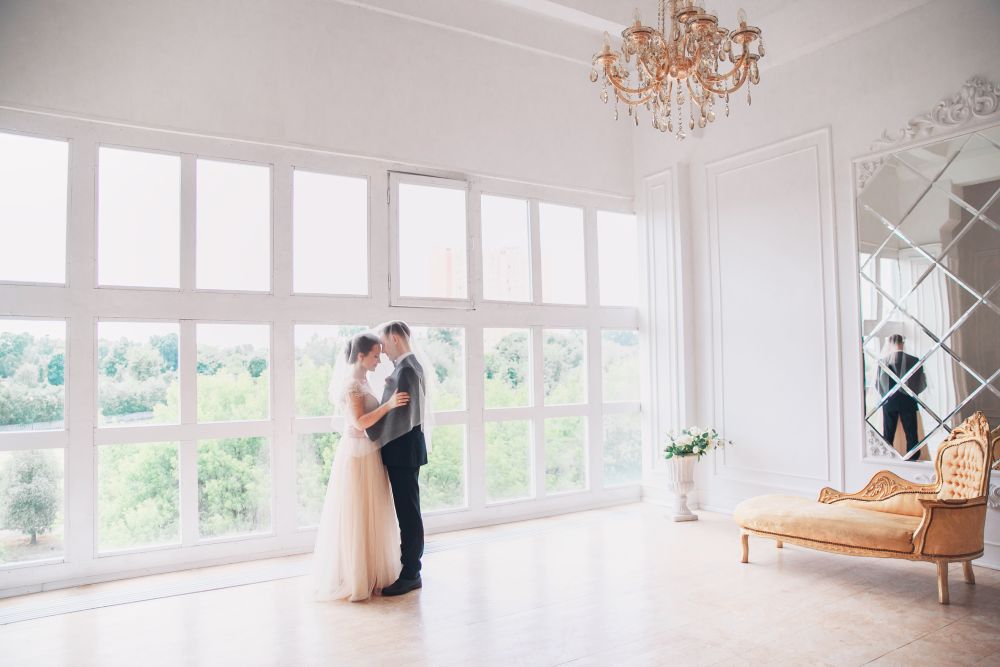 Ein Paar führt den Hochzeitstanz in einem hellen, eleganten Raum mit großen Gitterfenstern auf, die den Blick auf das üppige Grün draußen freigeben. Ein goldener Kronleuchter hängt über der Decke und eine verspiegelte Wand fängt die Spiegelung der Szene ein. In der Nähe steht ein dekoratives Sofa anmutig am Fenster.