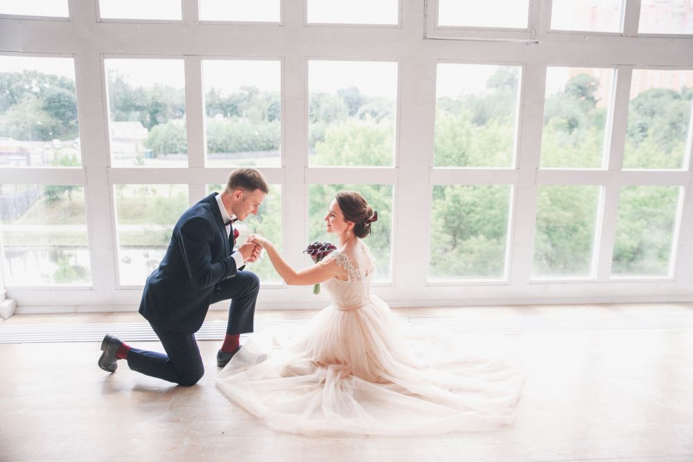 Ein Mann kniet vor einer Frau in einem leichten, fließenden Kleid und hält ihre Hand. Sie befinden sich in einem hellen Raum mit großen Fenstern, die einen grünen, malerischen Blick nach draußen bieten. Die Szene wirkt magisch, als würde der Hochzeitstanz gleich beginnen, während die Frau einen Strauß dunkler Blumen umklammert.