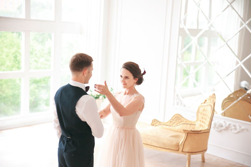 In einem hellen, eleganten Raum mit großen Fenstern und einer verspiegelten Wand steht ein Paar und lächelt sich in Vorfreude auf den Hochzeitstanz an. Die Frau trägt ein hellrosa Kleid und der Mann eine schwarze Weste über seinem weißen Hemd. Ein reich verziertes Sofa rundet die Szene ab.