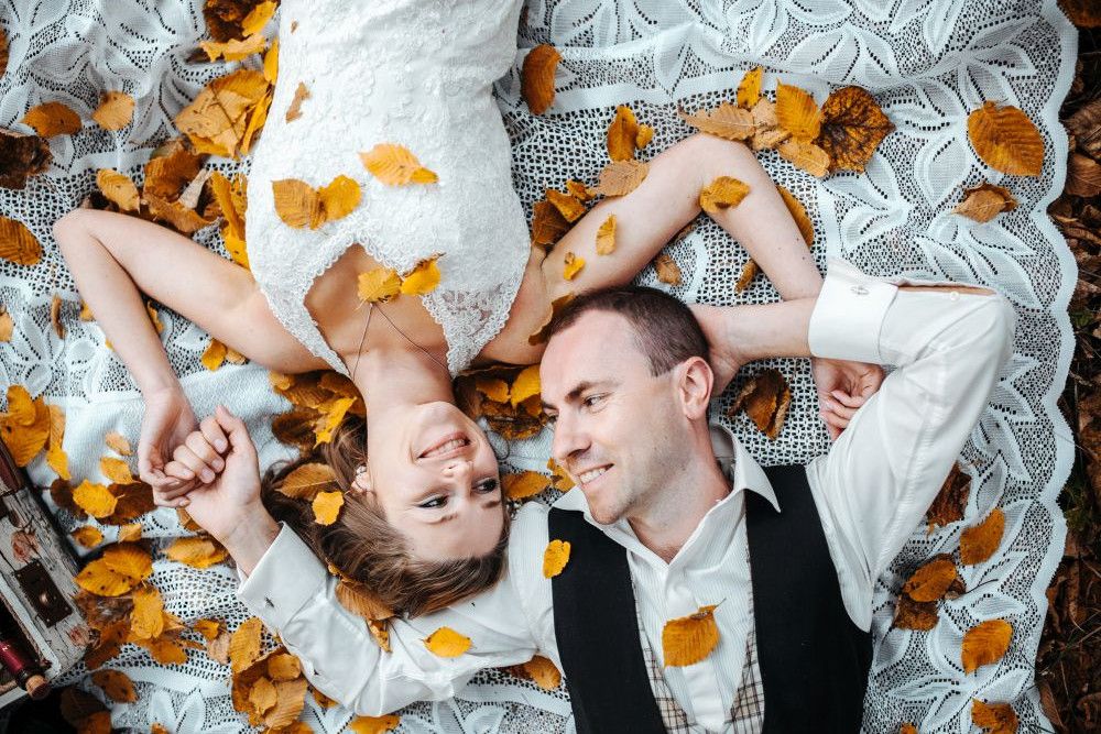 Ein Paar liegt auf einer weißen Spitzendecke, die mit orangefarbenen Blättern bedeckt ist. Die Frau im weißen Kleid liegt lächelnd auf dem Rücken und hält die Hand des Mannes neben ihr, der ein weißes Hemd und eine dunkle Weste trägt. Sie sind von verstreuten Blättern umgeben.