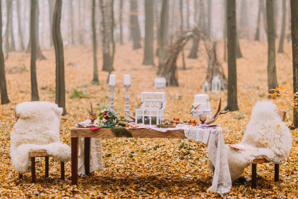 Ein rustikal gedeckter Tisch für ein Abendessen in einem Herbstwald mit zwei pelzbezogenen Stühlen. Der Tisch ist mit Kerzen, einem Blumenarrangement und einem Kuchen geschmückt. Gefallene Blätter bedecken den Boden und im Hintergrund ist ein dekorativer Holzbogen zu sehen.