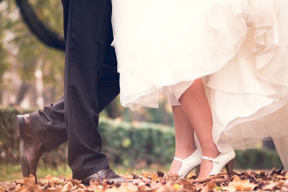 Braut und Bräutigam stehen dicht beieinander, von der Hüfte abwärts sichtbar. Der Bräutigam trägt dunkle Hosen und Schuhe, während die Braut ein weißes Kleid mit hohen Absätzen trägt. Sie sind von gefallenen Blättern auf dem Boden umgeben.