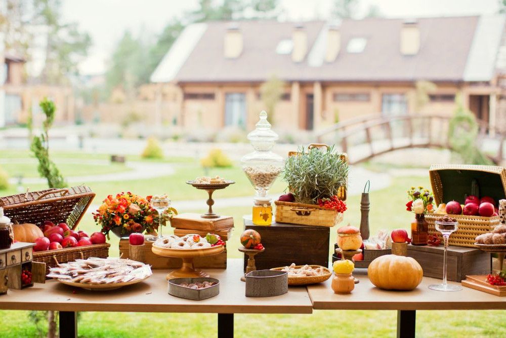Im Freien steht ein Tisch mit verschiedenen Speisen und Dekorationen, darunter Obstkörbe, Gebäck auf Tellern, ein Glasgefäß mit Deckel und ein kleiner Kürbis. Im Hintergrund sind Grünpflanzen und eine Holzbrücke zu sehen.