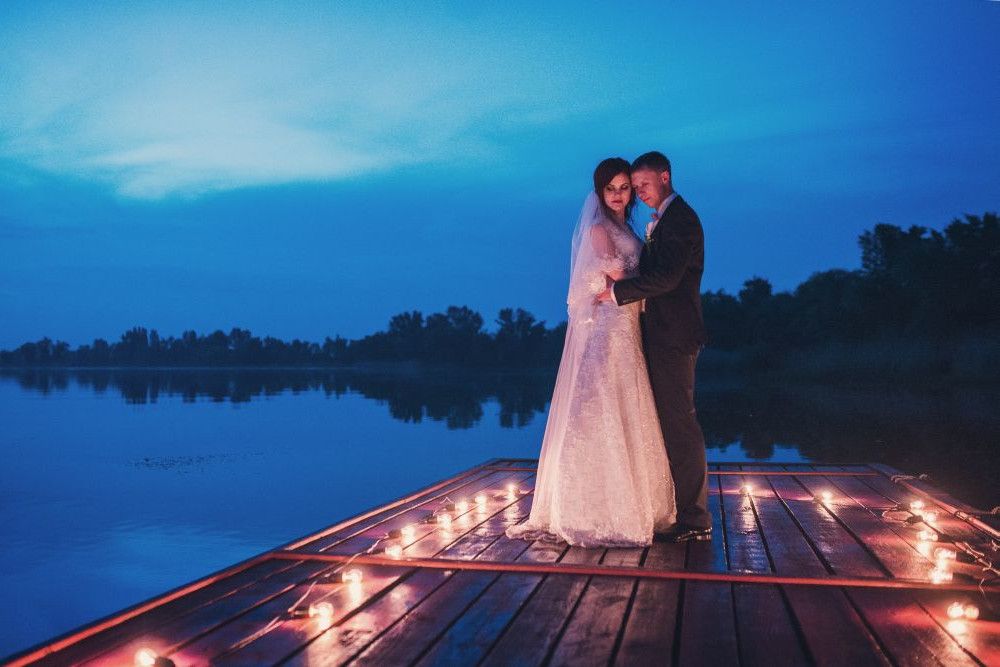 Ein Paar in Hochzeitskleidung steht auf einem Holzsteg, umgeben von brennenden Kerzen. Die Szene spielt in der Abenddämmerung mit einem ruhigen See und einem blauen Himmel im Hintergrund. Das Paar umarmt sich und blickt in die Kamera.