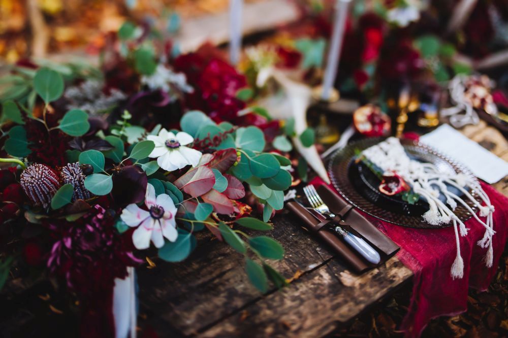 Ein rustikaler Holztisch ist mit einem üppigen Blumenarrangement aus roten und weißen Blumen und grünem Laub geschmückt. Es gibt Gedecke mit Besteck, einem Teller und einer roten Serviette. Kerzen und dekorative Elemente runden das Arrangement ab.