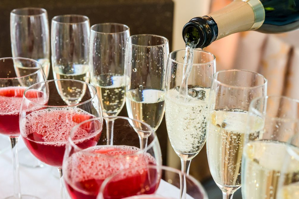 Es werden mehrere Gläser mit roten und weißen prickelnden Getränken ausgestellt. Eine Flasche Champagner wird in eines der hohen Gläser gegossen, wodurch Blasen entstehen. Die Kulisse suggeriert eine festliche oder Party-Atmosphäre.