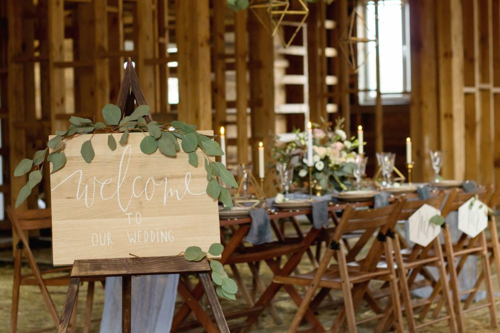 Eine rustikale Hochzeitsdekoration in einer Holzscheune zeigt ein Willkommensschild auf einer mit Grünpflanzen geschmückten Staffelei. Im Hintergrund steht ein langer Tisch mit Kerzen und Blumenarrangements, umgeben von Holzstühlen mit blauen Stoffakzenten.