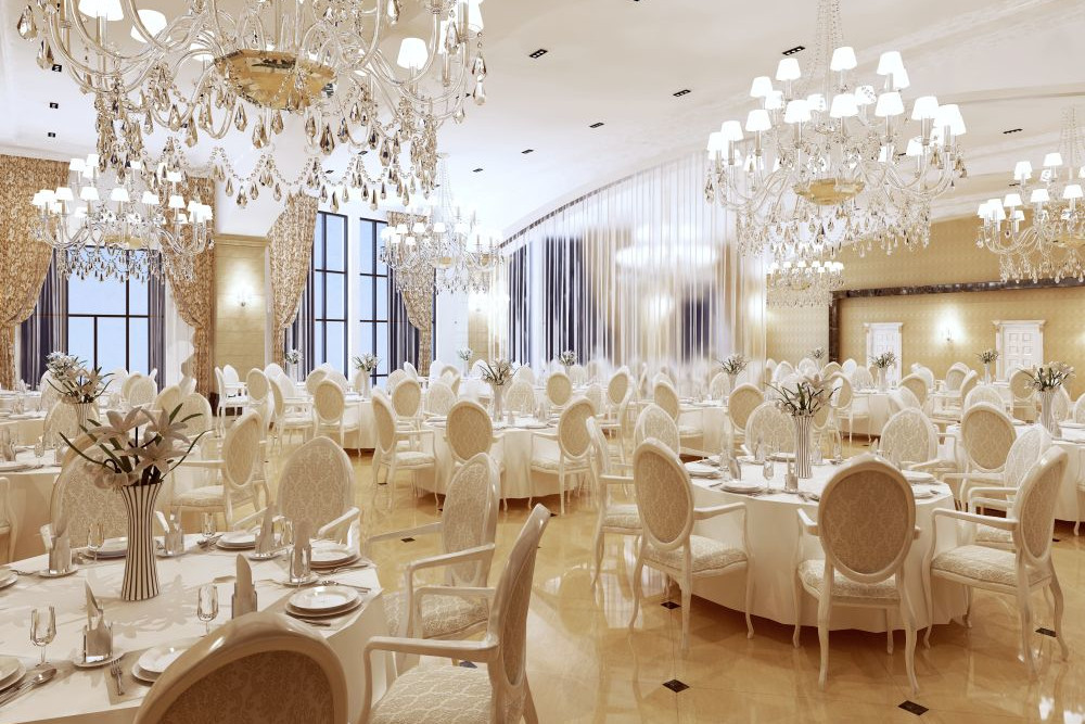 Ein geräumiger, eleganter Bankettsaal, der mit mehreren Kronleuchtern an der Decke dekoriert ist. Der Raum verfügt über runde Tische mit weißen Tischdecken und Stühlen mit goldenen Akzenten. Große Fenster mit Vorhängen lassen den Raum mit natürlichem Licht durchfluten.