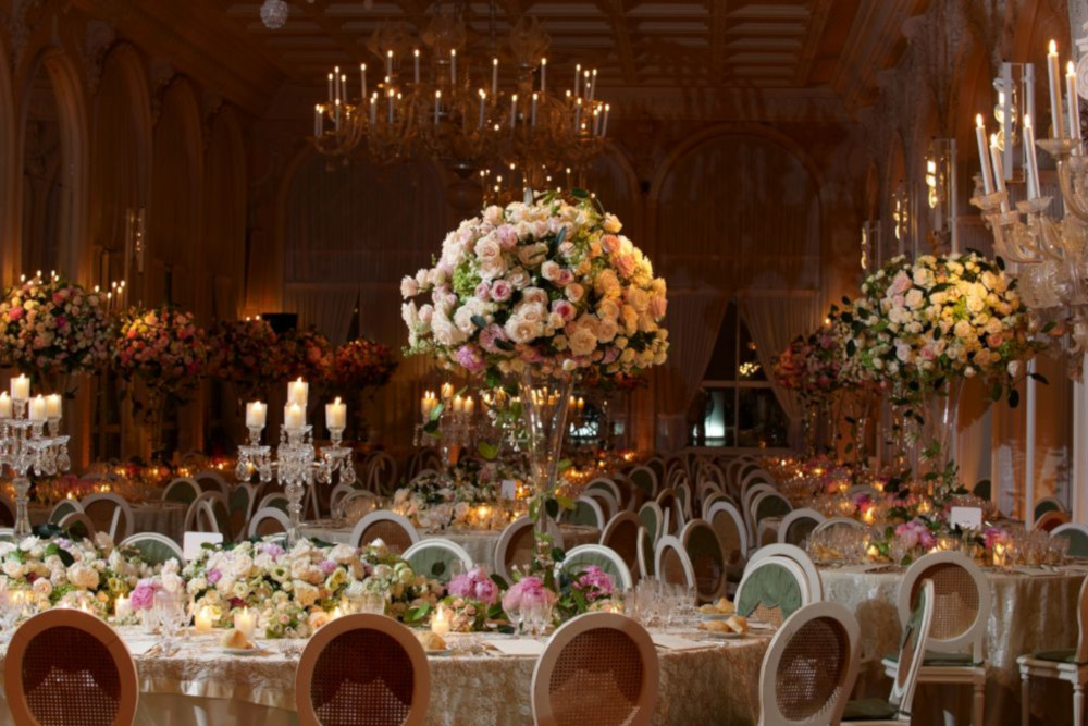 Ein eleganter Ballsaal ist für eine Veranstaltung großzügig dekoriert, mit kunstvollen Kronleuchtern, großen Blumengestecken mit Rosen und Tischen mit Kerzen und feinem Geschirr. Der Raum strahlt eine warme, luxuriöse Atmosphäre aus.