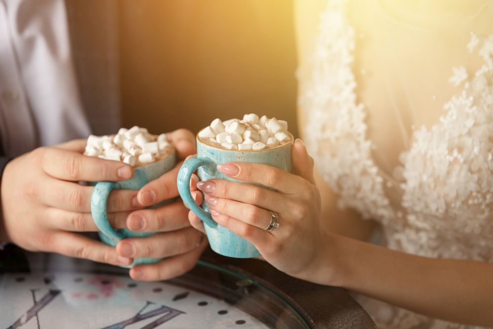 Zwei Personen halten blaue Tassen mit heißer Schokolade und Marshmallows. Eine Person trägt ein Spitzenkleid, die andere einen Anzug. Sie sitzen an einem Glastisch mit Uhrenmotiv. Sonnenlicht fällt hinein und schafft eine warme Atmosphäre.