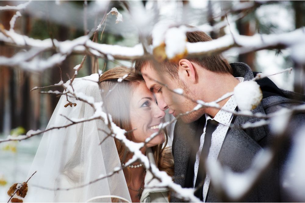 In einem malerischen verschneiten Wald teilen Braut und Bräutigam einen intimen Moment. Schneebedeckte Zweige rahmen ihre Gesichter ein, während sie einander anschauen. Mit der Braut im Schleier und dem Bräutigam im Anzug fängt das Bild den Zauber von „Heiraten im Winter“ perfekt ein.