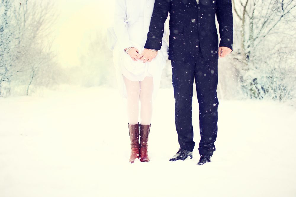 Ein Paar steht händchenhaltend im Schnee, der teilweise von der Hüfte abwärts sichtbar ist. Die Person links trägt einen weißen Mantel und braune Stiefel, während die Person rechts einen dunklen Anzug und schwarze Schuhe trägt. Um sie herum fällt leichter Schnee.