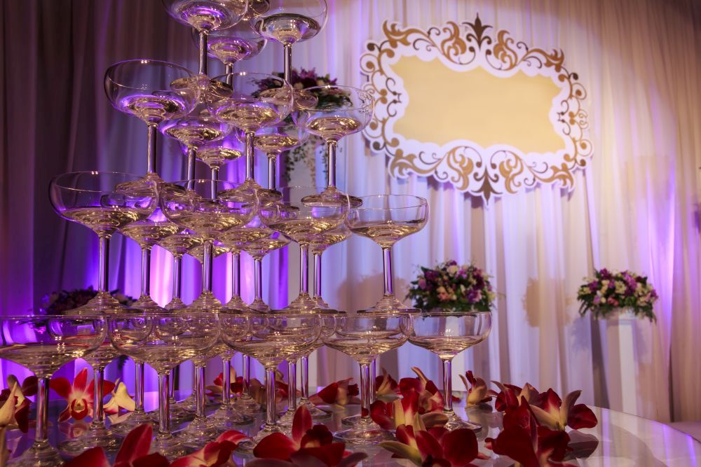 Auf einem Tisch steht ein mehrstöckiger Champagnerglasturm, umgeben von roten und weißen Blütenblättern. Im Hintergrund sind dekorative weiße Vorhänge, violette Beleuchtung, Blumenarrangements und ein leeres, verziertes goldenes Schild zu sehen.