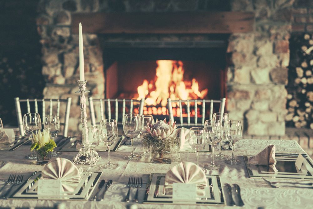 Ein Esstisch ist elegant mit Gläsern, Besteck und gefalteten Servietten gedeckt und steht vor einem Steinkamin mit brennendem Feuer. Chiavari-Stühle umgeben den Tisch, der mit einem Blumengesteck und Kerzenständern dekoriert ist.