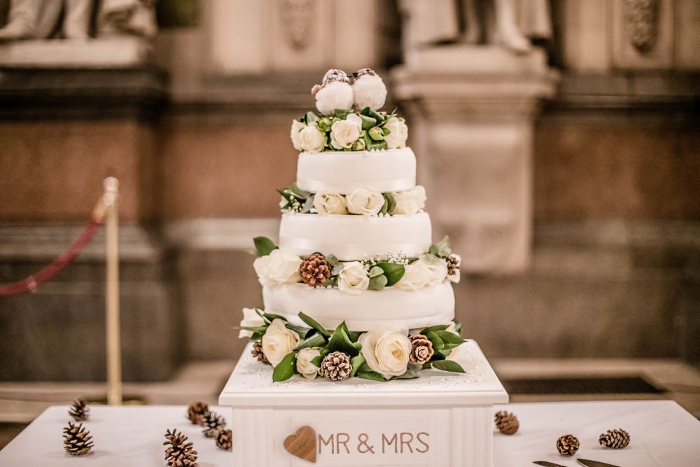 Eine vierstöckige weiße Hochzeitstorte, geschmückt mit weißen Rosen, Tannenzapfen und Laub. Der Tortenaufsatz zeigt zwei kleine Figuren. Die Torte wird auf einem Ständer mit einer „Mr & Mrs“-Plakette präsentiert, umgeben von weiteren Tannenzapfen.