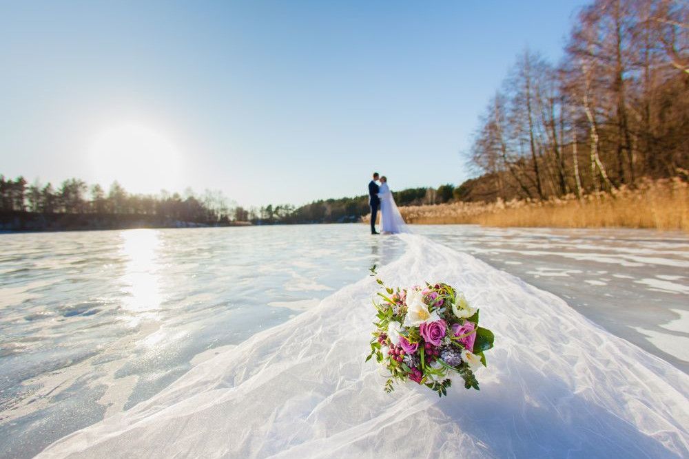 Braut und Bräutigam stehen umarmend auf einem zugefrorenen See unter einem klaren blauen Himmel. Im Vordergrund ist ein Strauß rosa und weißer Blumen auf der langen Schleppe des Brautkleides zu sehen, die über das Eis ausgebreitet ist. Bäume säumen den Horizont.