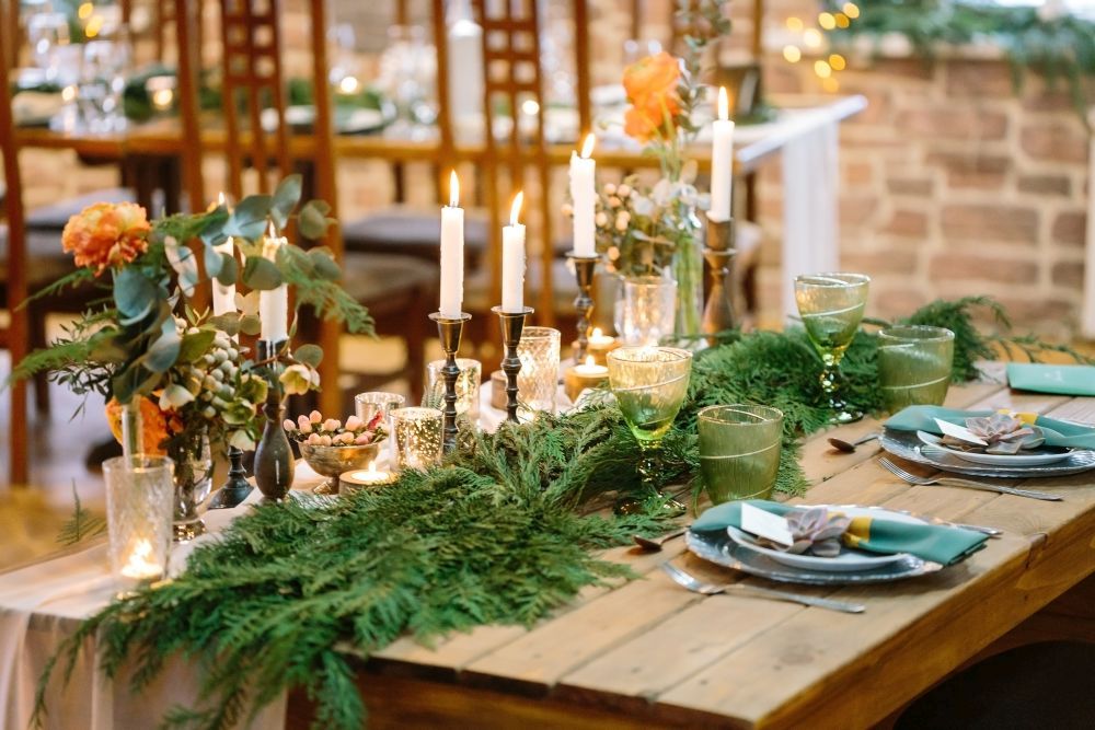 Ein hölzerner Esstisch ist mit grünen Servietten und Glaswaren gedeckt. Ein Tafelaufsatz aus Grünpflanzen, Kerzen und orangefarbenen Blumen schmückt den Tisch. Im Hintergrund sind Holzstühle und eine Backsteinmauer zu sehen.