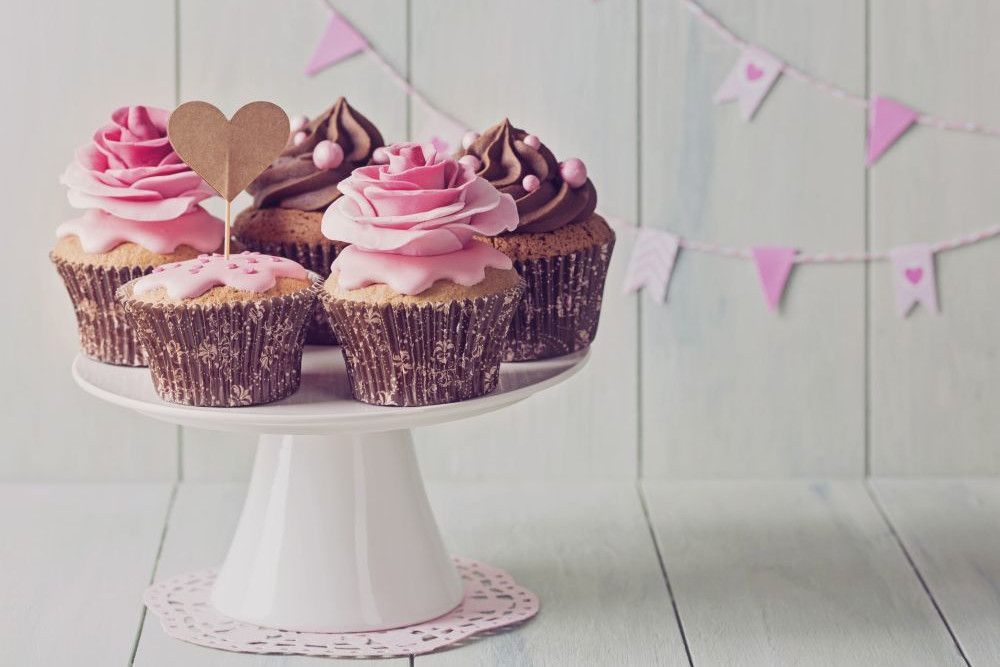 Eine entzückende Auswahl an Cupcakes mit rosa und schokoladenfarbenem Zuckerguss ziert einen weißen Tortenständer. Ein Cupcake hat einen herzförmigen Aufsatz und weckt den Charme von „Heirate im Frühling“. Im Hintergrund schmücken rosa und weiße Wimpel eine helle Holzwand.