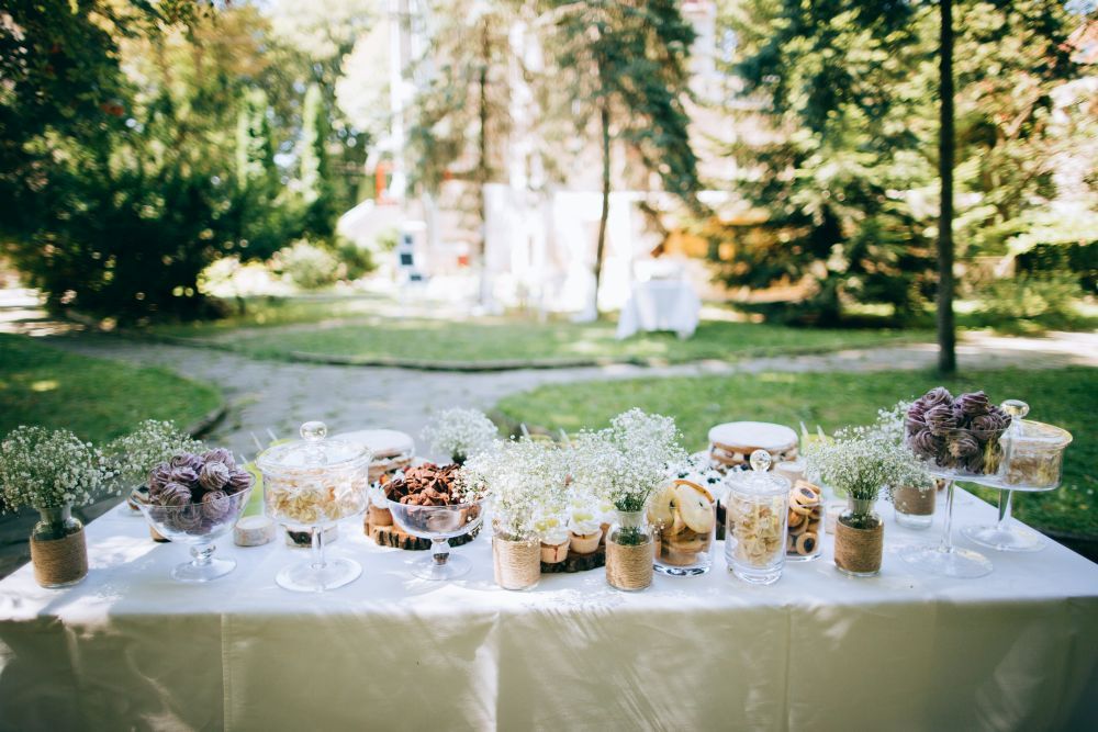 Ein wunderschön arrangierter Desserttisch im Freien vor einer üppig grünen Gartenkulisse fängt die Essenz von „Heirate im Frühling“ ein. Der Tisch bietet Süßigkeiten in eleganten Gläsern und Ständern, ergänzt durch kleine weiße Blumenarrangements für eine einladende und romantische Präsentation.
