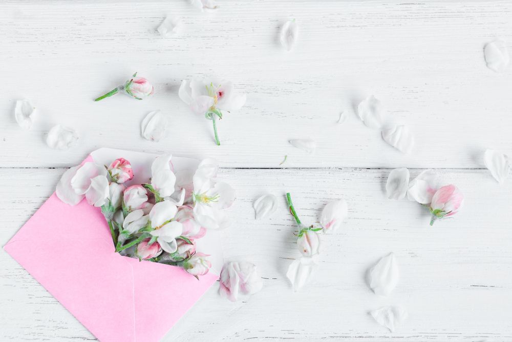 Ein rosa Umschlag auf einer weißen Holzoberfläche ist mit zarten weißen und rosa Blumen gefüllt und fängt die Essenz von „Heiraten im Frühling“ ein. Verstreute Blütenblätter um den Umschlag herum sorgen für eine sanfte, romantische Szene.