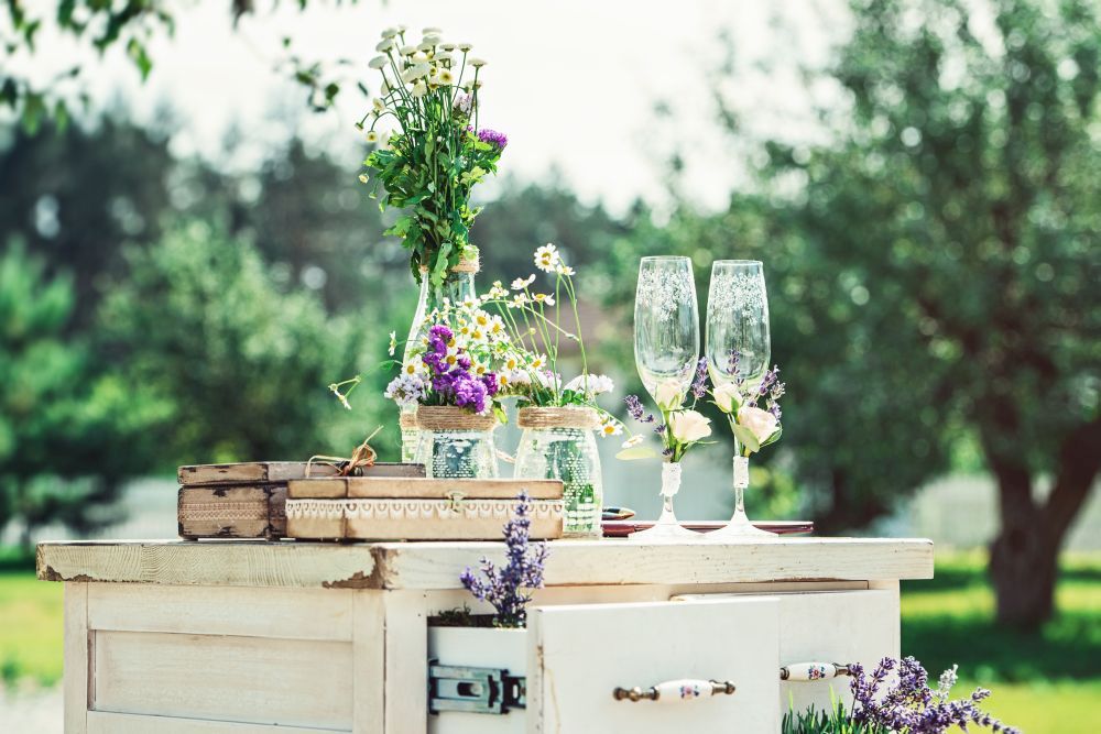 Ein rustikaler Tisch im Freien, dekoriert mit Wildblumen in Glasgefäßen, Champagnergläsern und Vintage-Boxen, bietet die perfekte Kulisse für „Heirate im Frühling“. Die Kulisse ist ein üppiger Garten, der eine ruhige und einladende Atmosphäre schafft.