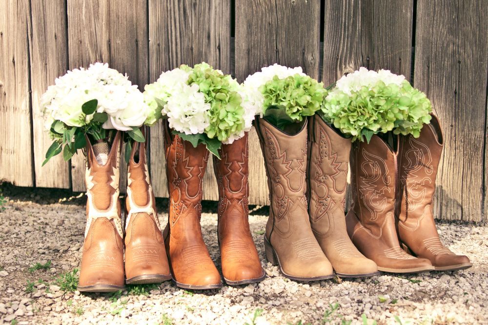 Eine Reihe Cowboystiefel, aufgereiht unter einer Holzwand, jeder mit weißen und grünen Hortensienblüten geschmückt, ergibt eine bezaubernde Szene, die an „Heiraten im Frühling“ erinnert, und ruht wunderschön auf einer Kiesfläche.