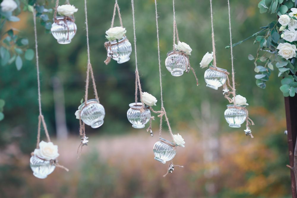 Hängende Glasgefäße mit weißen Blumen und Schnüren ergeben vor einem unscharfen Hintergrund aus grünem Laub eine romantische Dekoration, perfekt zum Heiraten im Frühling.