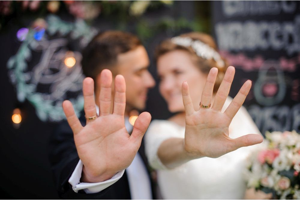 Braut und Bräutigam strecken ihre Hände in die Kamera und präsentieren ihre Eheringe. Leicht unscharf fangen sie die Essenz von „Heiraten im Frühling“ ein, wobei die Braut einen Blumenstrauß vor einem dekorativen Hintergrund hält, der für ihren besonderen Tag sanft beleuchtet ist.