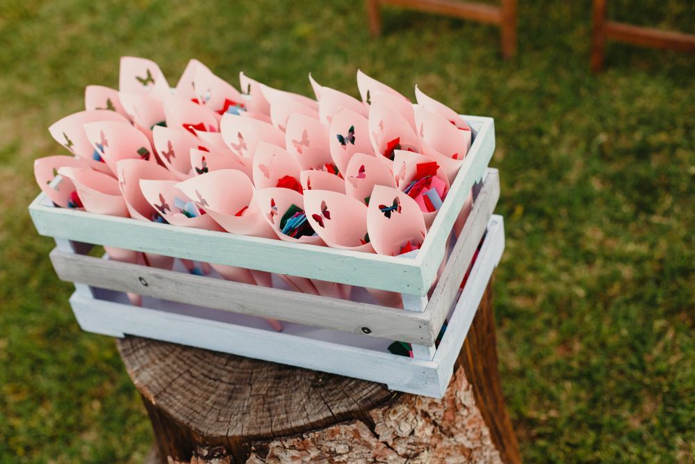 Eine Holzkiste enthält zahlreiche rosa Papiertüten mit herzförmigen Ausschnitten, perfekt für „Heiraten im Frühling“. Die mit buntem Konfetti gefüllte Kiste steht elegant auf einem Baumstumpf vor einem Hintergrund aus saftigem grünem Gras.