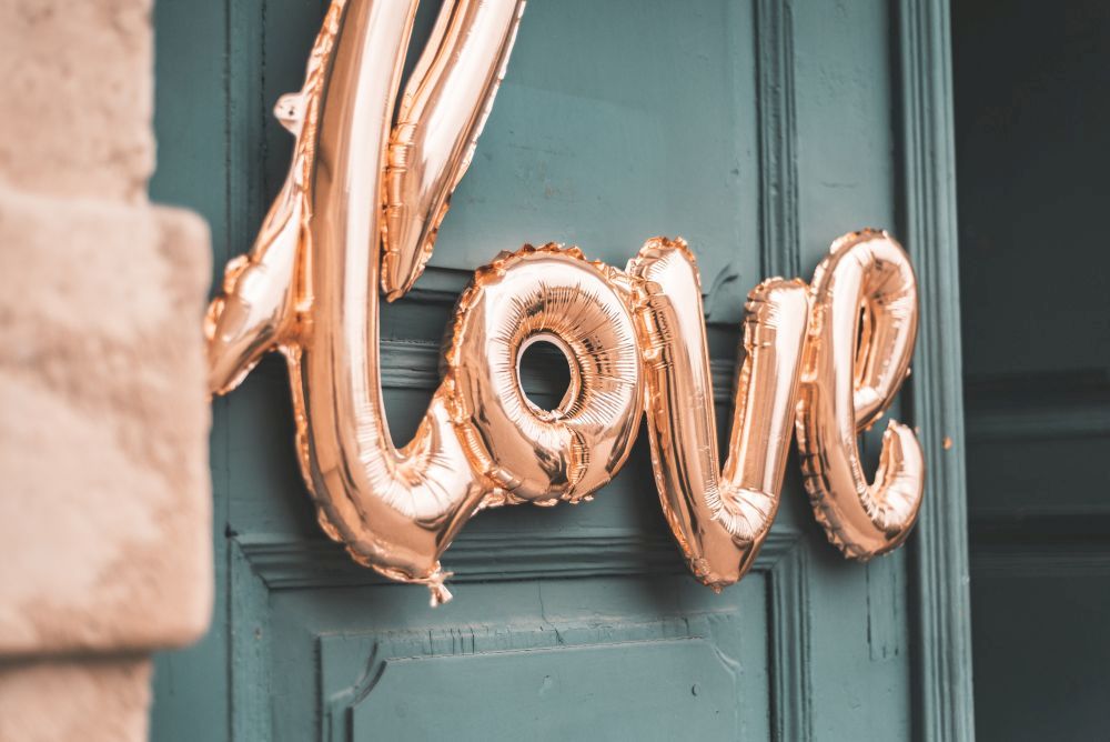 Vor einer blaugrünen Tür prangen goldene Folienballons mit der Aufschrift „Love“, die dem eleganten Glanz der Ballons einen rustikalen Charme verleihen – perfekt für alle, die „Heiraten im Frühling“ planen.