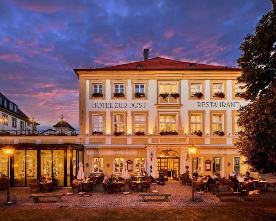 Ein dreistöckiges Gebäude mit einem Schild mit der Aufschrift „Hotel zur Post Altötting“ steht vor einem violett-orangefarbenen Abendhimmel. Im Außenbereich wimmelt es von Menschen, warmes Licht erhellt die Fassade. Auf der rechten Seite steht ein großer Baum.