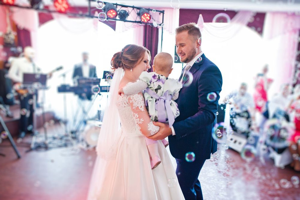 Braut und Bräutigam tanzen bei einer Hochzeitsfeier mit einem Baby im Arm. Im Hintergrund sind sie von Seifenblasen und Gästen umgeben. Man sieht eine Band, die Musik spielt, und der Raum ist mit weißen Vorhängen und festlichen Lichtern geschmückt.