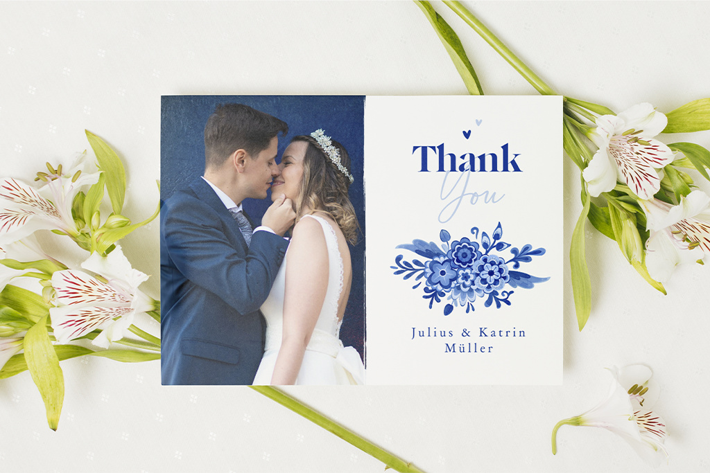 Eine Dankeskarte zur Hochzeit zeigt ein Paar, das einen intimen Moment miteinander teilt. Auf der Karte steht „Danke“ mit Blumenschmuck und den Namen „Julius & Katrin Müller“. Weiße Blumen rahmen die Karte auf hellem Hintergrund ein.