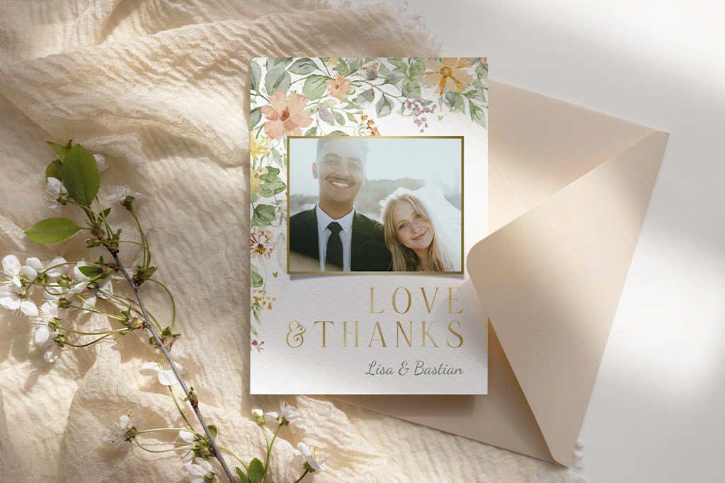 Eine Dankeskarte mit Blumenrand und einem Foto eines lächelnden Paares. Der Text lautet „LOVE & THANKS“ gefolgt von „Lisa & Bastian“. Die Karte liegt auf einem strukturierten Stoff neben einem offenen Umschlag und einigen Zweigen weißer Blüten.