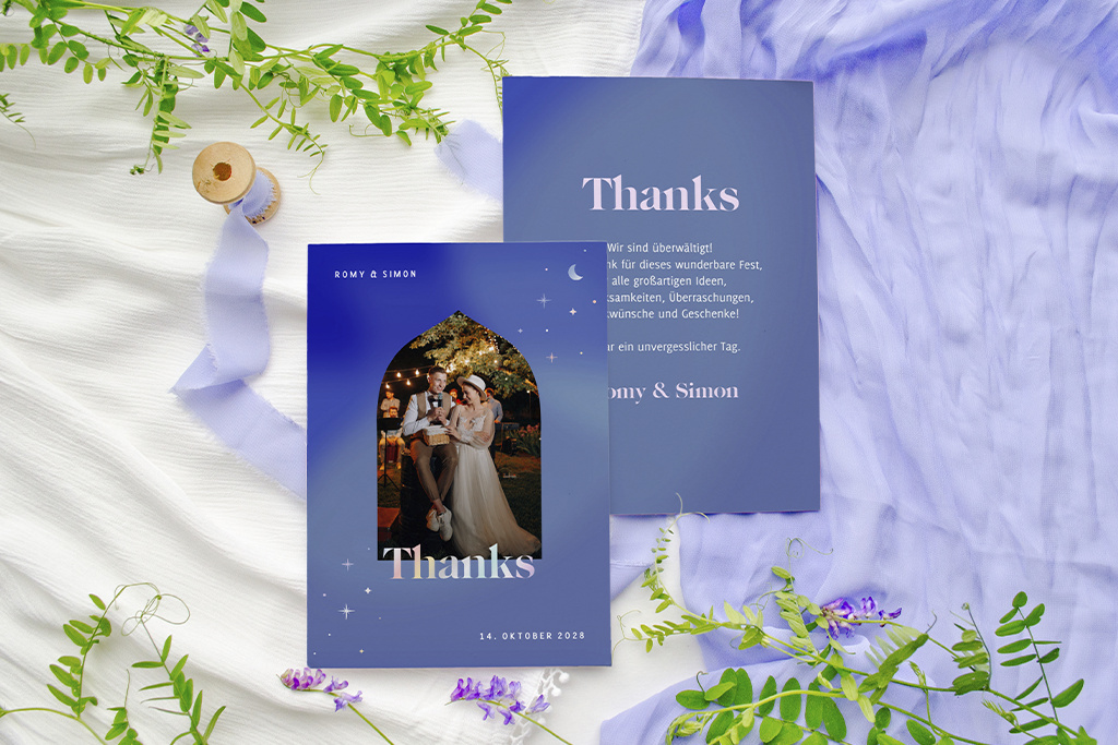 Eine Dankeskarte mit einem frisch verheirateten Paar liegt auf einer strukturierten Oberfläche mit Lavendelzweigen und grünen Blättern. Das Kartendesign umfasst Pastellfarben und einen eleganten Text, daneben liegt eine Spule mit Band.