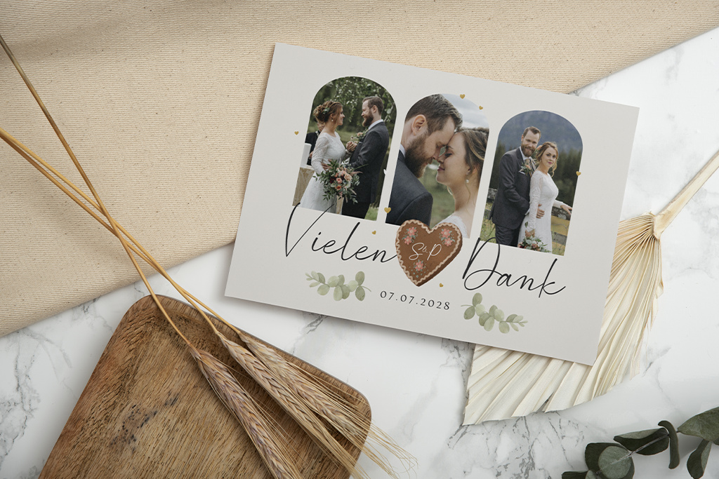 Eine Dankeskarte zur Hochzeit mit drei Fotos eines Paares. Auf der Karte steht „Vielen Dank“ mit einem Herz und Initialen in der Mitte, datiert 07.07.2023. Sie ruht auf einer Marmoroberfläche mit Dekorationen aus Holz und getrockneten Pflanzen.