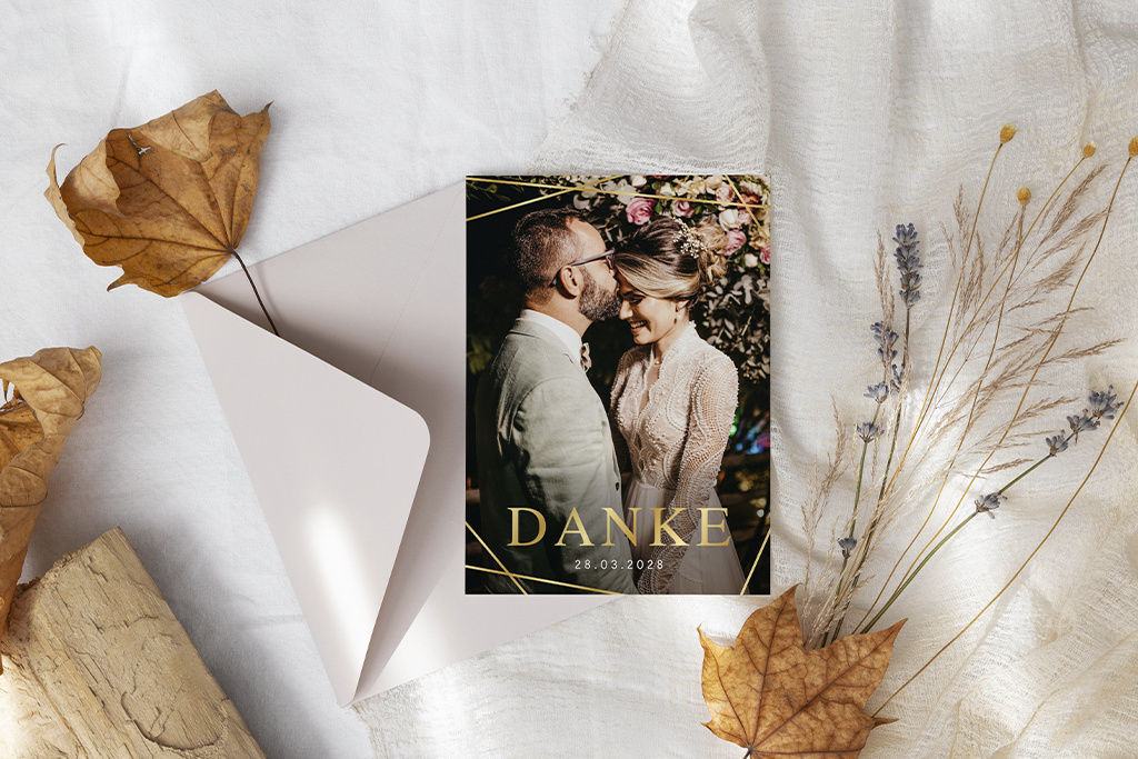 Danksagungskarte mit einem Foto eines lächelnden Paares auf einem weißen Tuch, daneben ein offener Umschlag und getrocknete Blätter.
