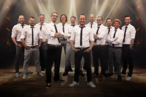 Eine Gruppe von zwölf Männern steht auf einer Bühne mit dramatischer Beleuchtung im Hintergrund. Sie tragen weiße Hemden und schwarze Krawatten. Die meisten von ihnen haben die Arme verschränkt, einige haben die Hände in den Taschen.