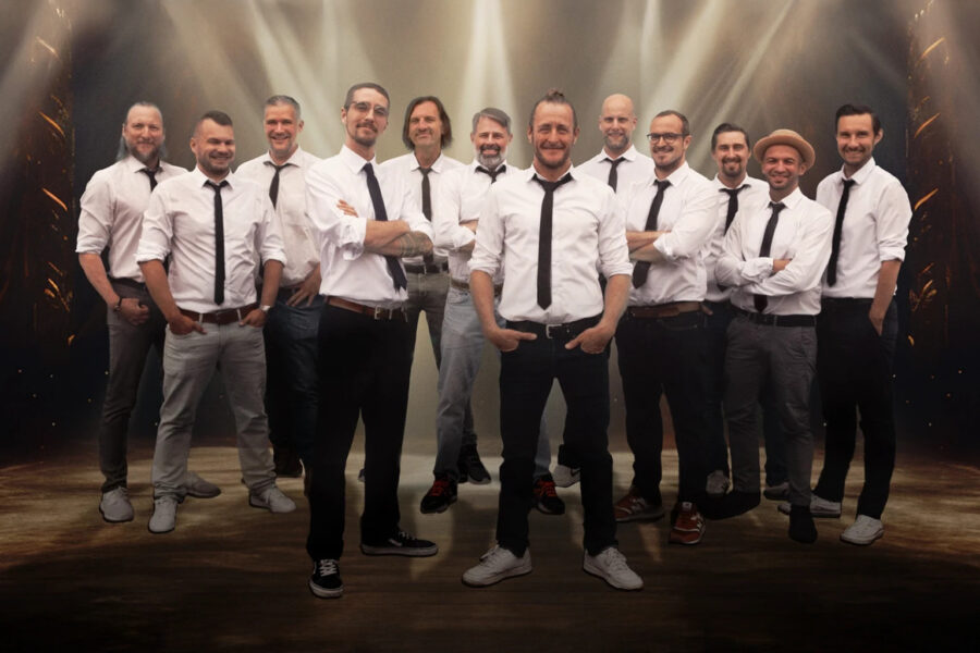 Eine Gruppe von zwölf Männern steht auf einer Bühne mit dramatischer Beleuchtung im Hintergrund. Sie tragen weiße Hemden und schwarze Krawatten. Die meisten von ihnen haben die Arme verschränkt, einige haben die Hände in den Taschen.