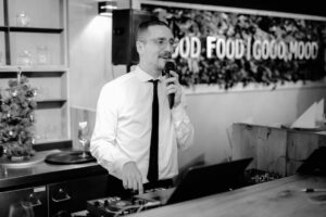 Ein Mann mit Brille und Krawatte spricht in ein Mikrofon, während er hinter einem DJ-Pult steht. Im Hintergrund ist eine Wand mit Grünpflanzen und den Worten „GUTES ESSEN, GUTE LAUNE“ zu sehen.