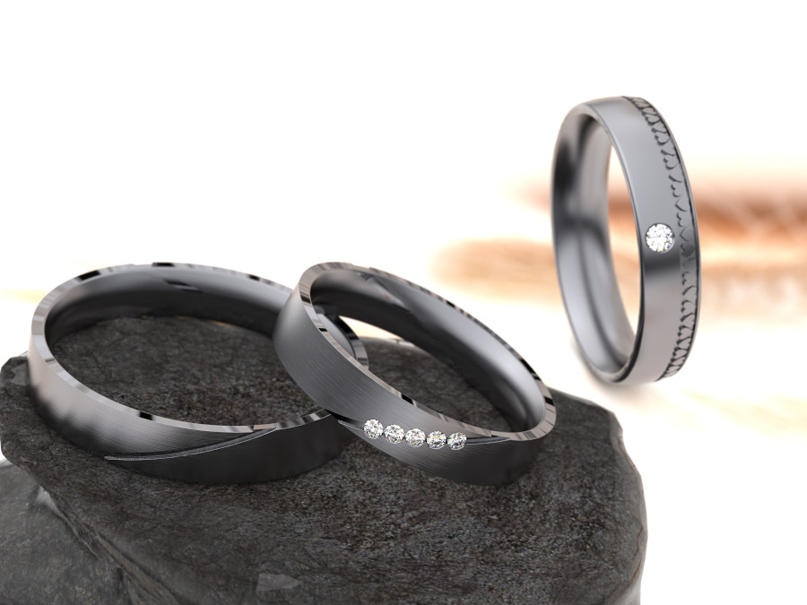 HOCHZEITde-Inspiration-Ratgeber-Aktuelles-Trauringe-aus-Tantal-vonjacob Drei elegante Ringe aus Tantal vor einem unscharfen Hintergrund. Zwei Ringe liegen auf einer Steinoberfläche und sind mit kleinen Edelsteinen verziert, während der dritte Ring aufrecht steht und einen einzelnen Edelstein aufweist.