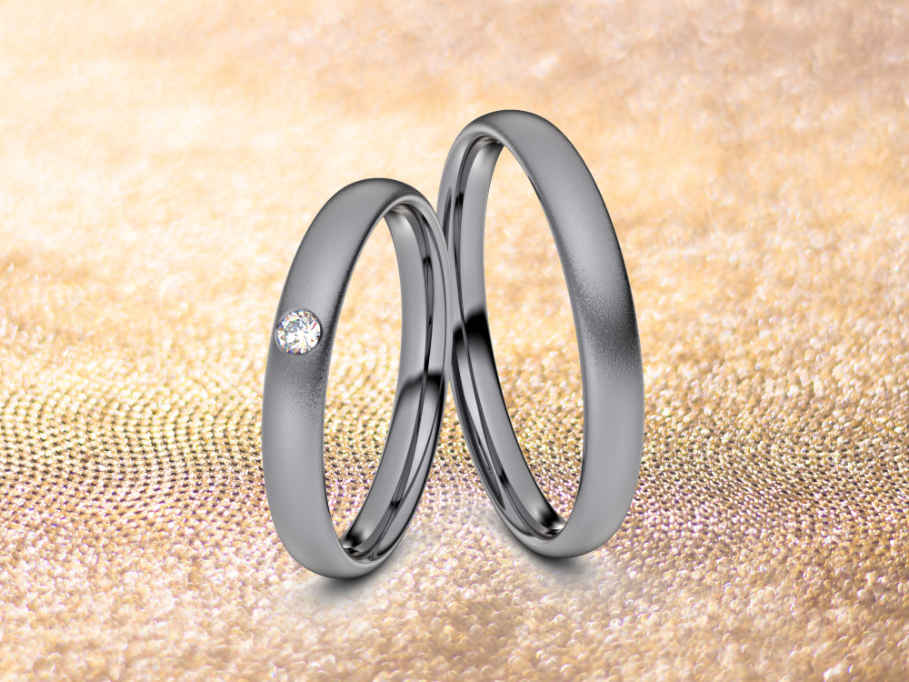 Zwei Ringe aus Tantal liegen nebeneinander auf einer strukturierten, goldenen Oberfläche. In den linken Ring ist ein kleiner Diamant eingearbeitet, während der rechte Ring schlicht ist. Der Hintergrund scheint sanft beleuchtet zu sein, um den metallischen Glanz zu verstärken.